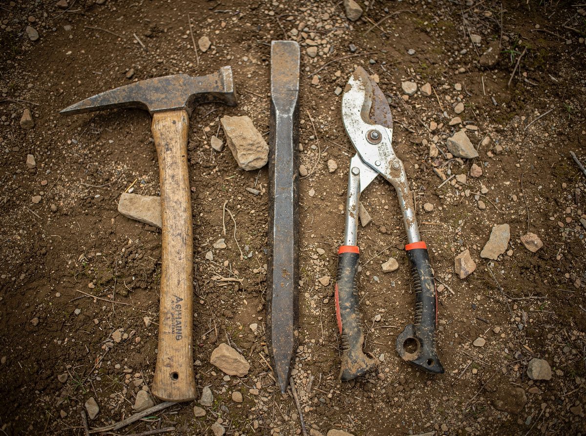 A mattock, a rock bar, and loppers laid out on packed dirt at a trail site